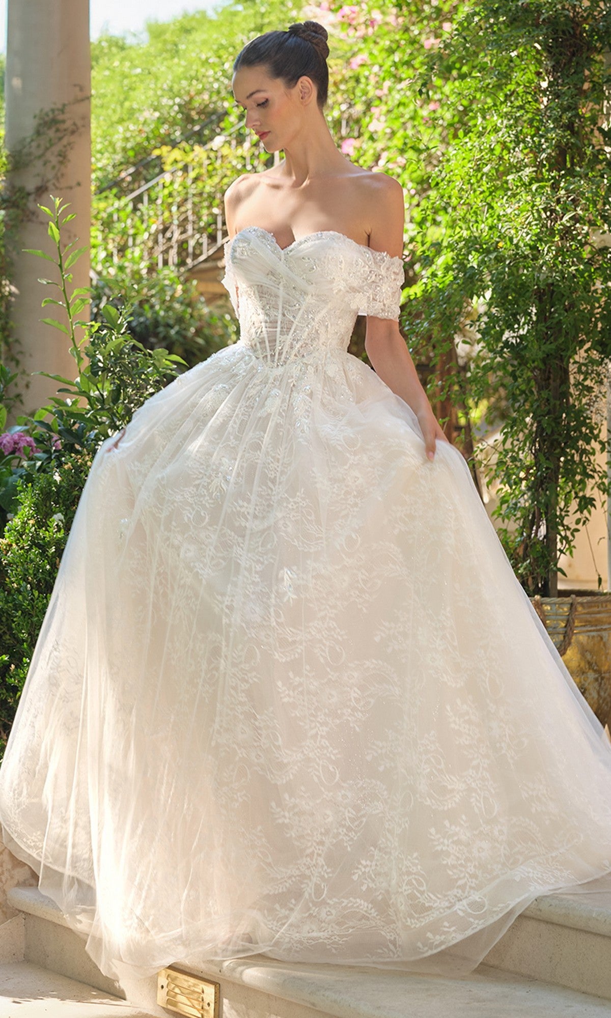 A woman stands outdoors in the Andrea and Leo WL090 long wedding dress, featuring an off-the-shoulder corset bodice and full lace skirt, surrounded by lush greenery.
